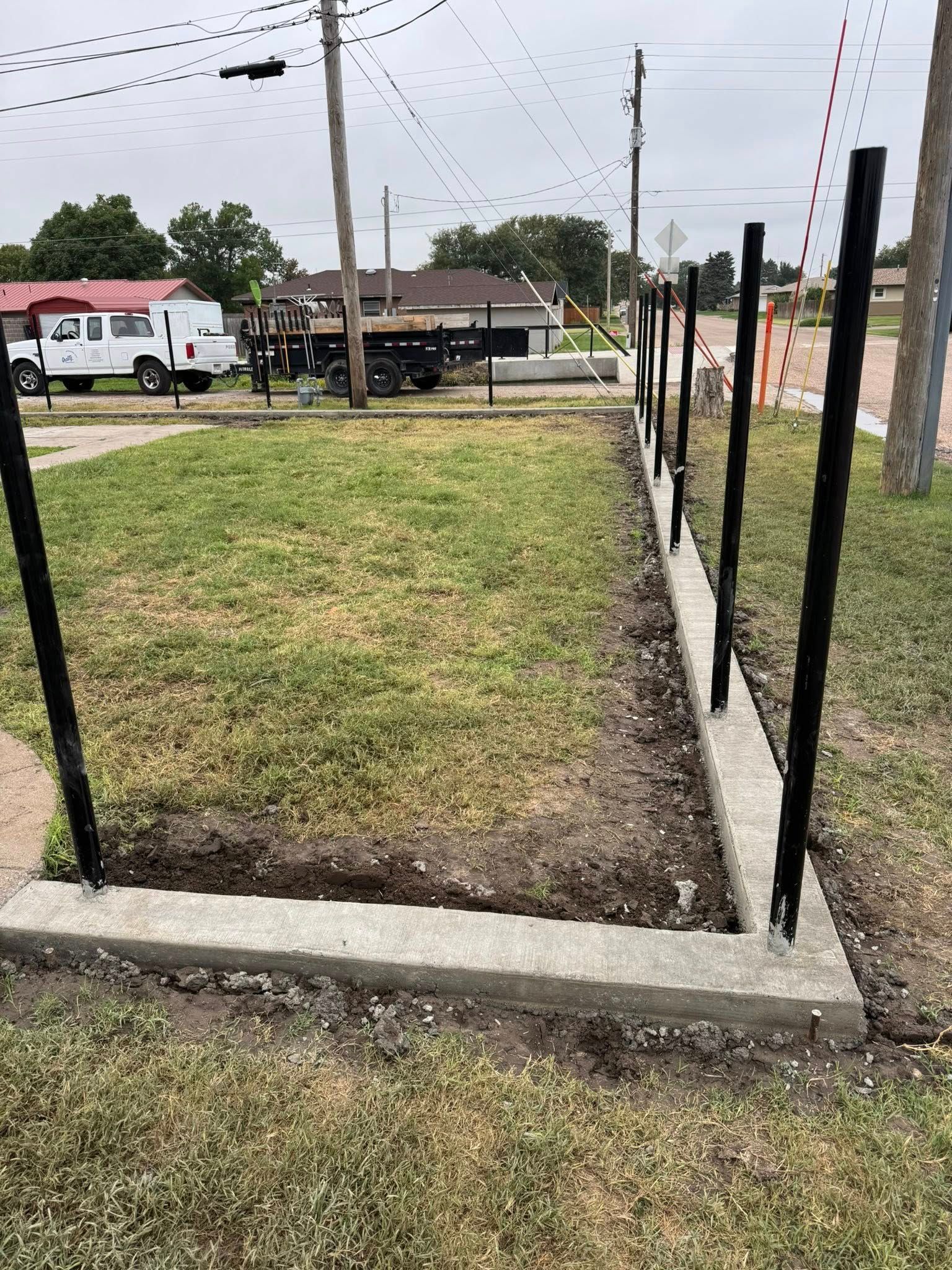 Small grassy lot bordered by concrete curbs and black posts beside a roadside under cloudy skies