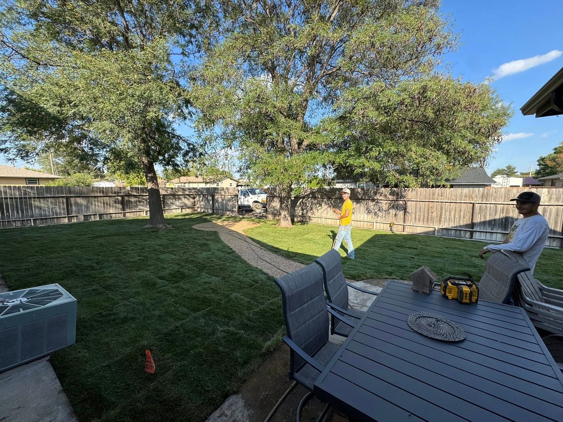 Backyard patio with a person and child near a table, trees, fence, and bright sunlight