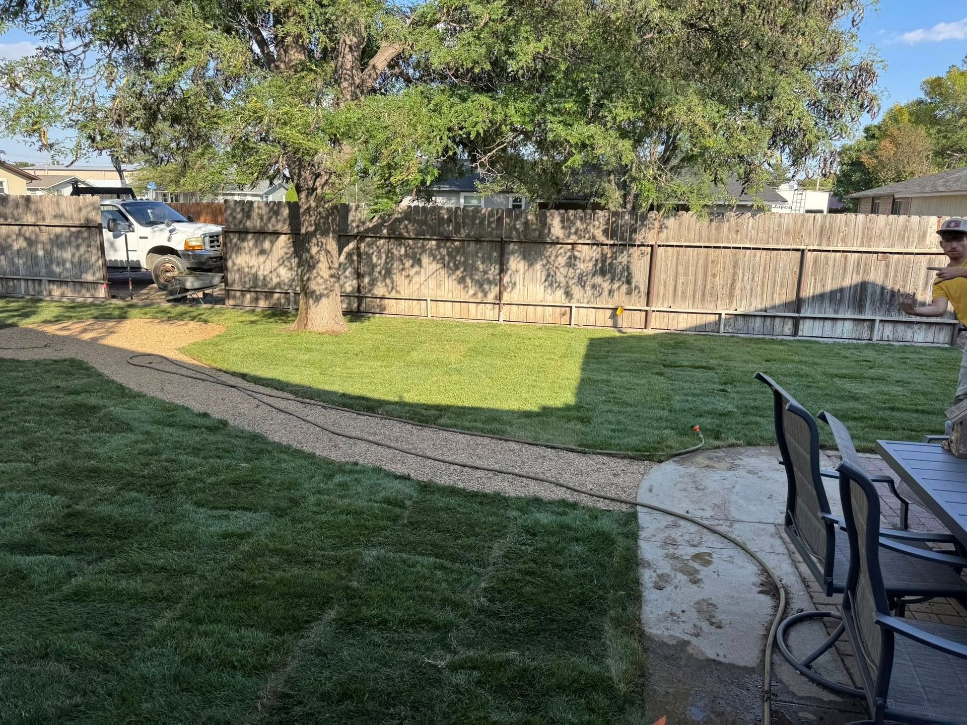 Backyard with green lawn, curved stone walkway, tree, fence, and patio chairs on the right.