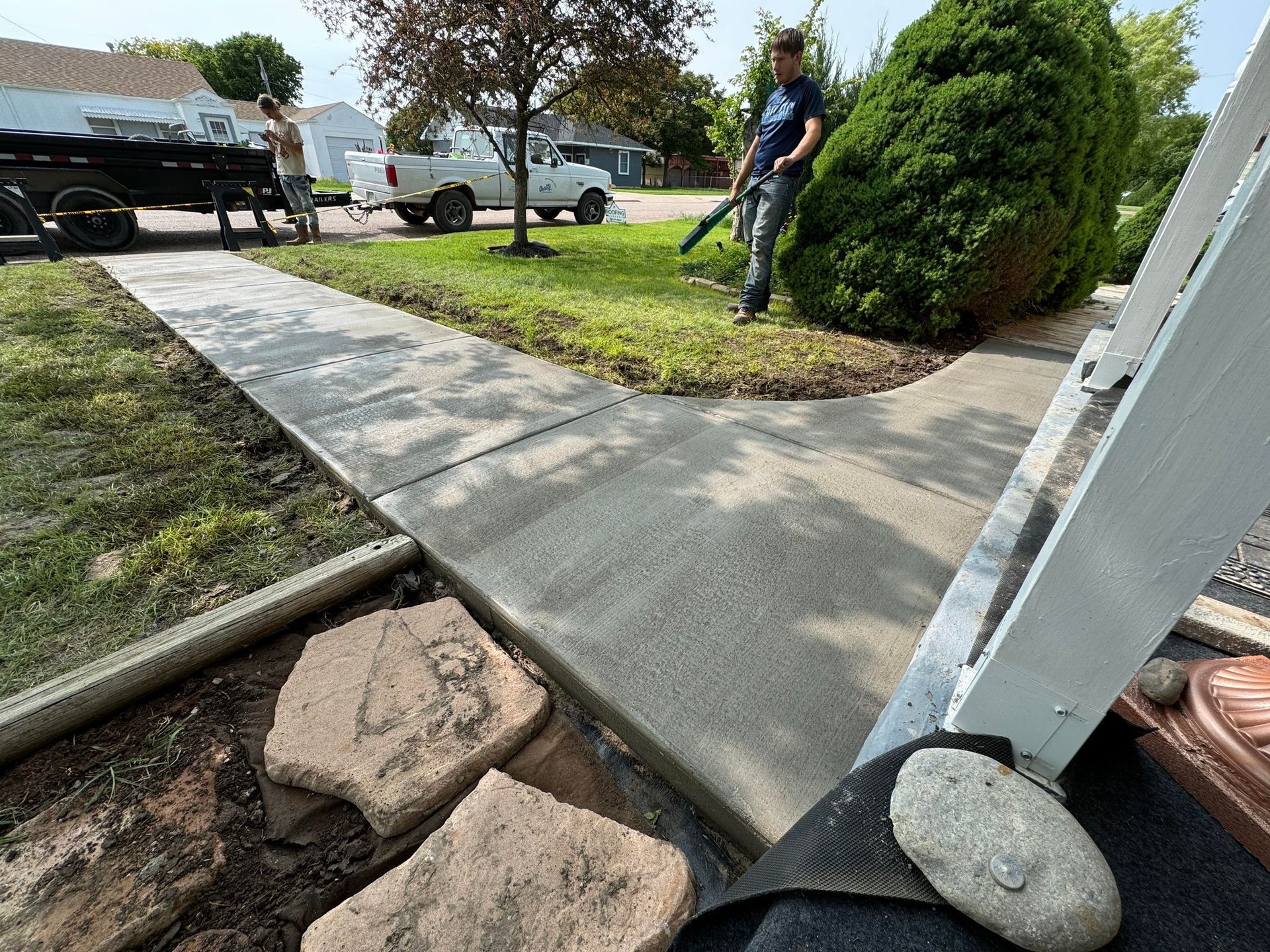 Sidewalk ramp with concrete path, rocks, and a person standing near a bush in a suburban yard