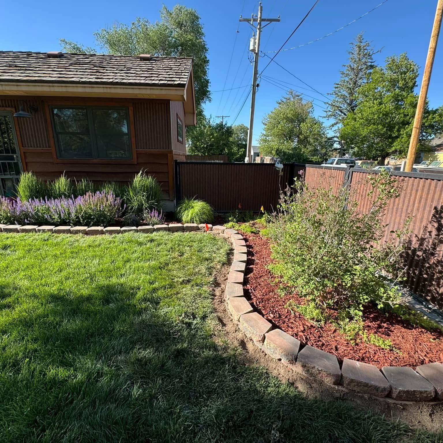 Backyard garden with curving stone border, green lawn, purple flowers, and a wooden shed under blue sky