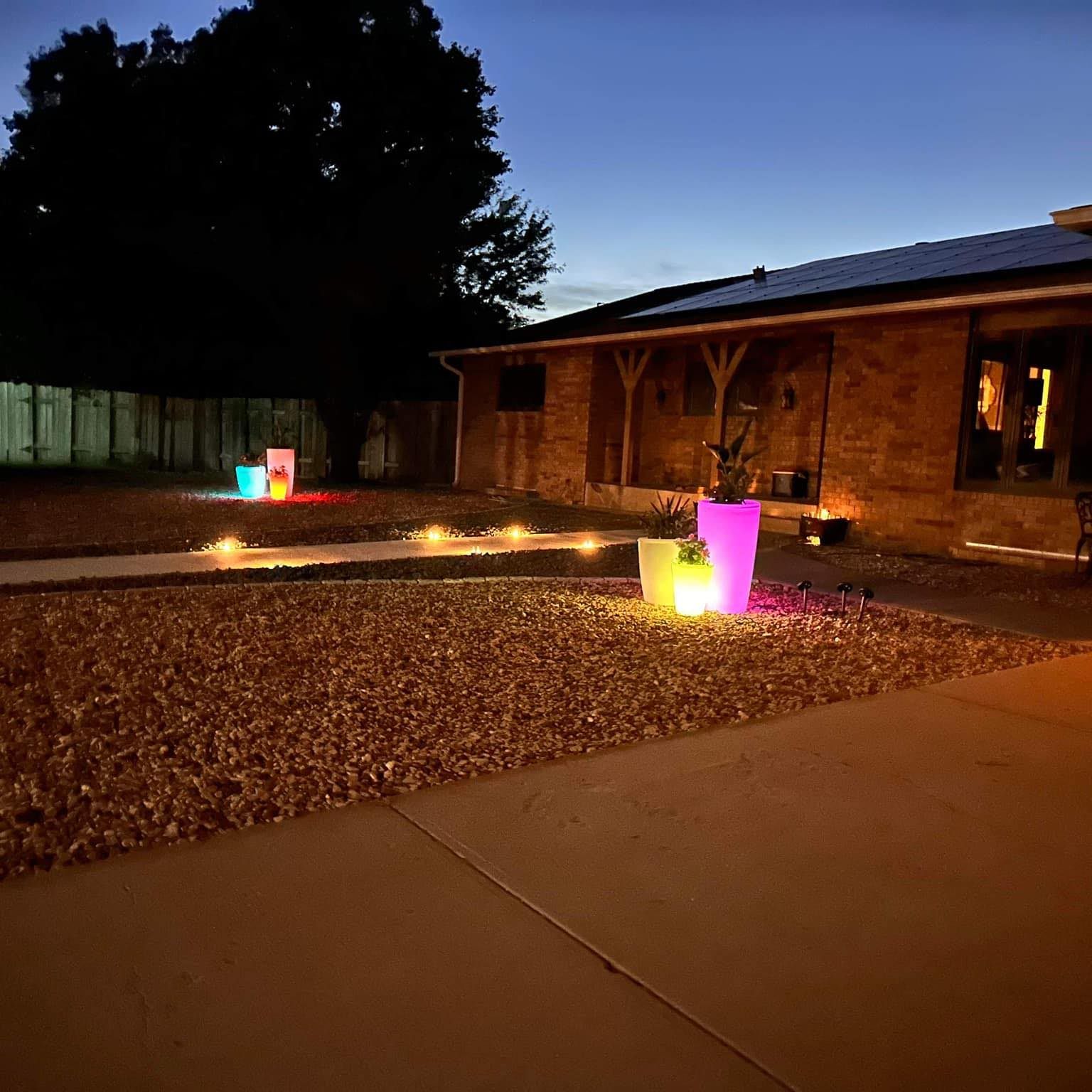 Front yard at dusk with glowing colorful light-up planters beside a brick house and walkway