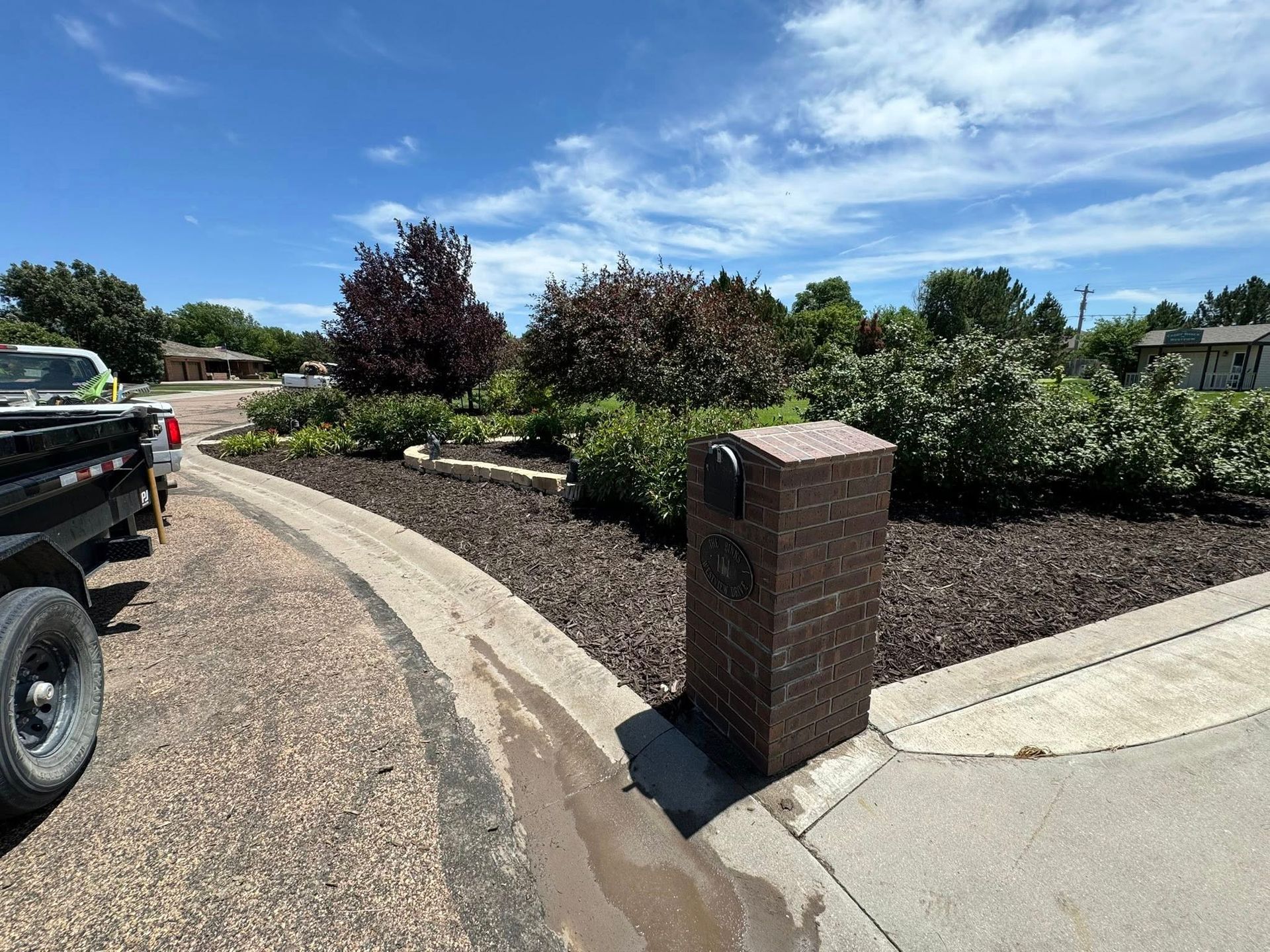 Residential landscape with a brick mailbox, curbed driveway, and shrubs under a blue sky