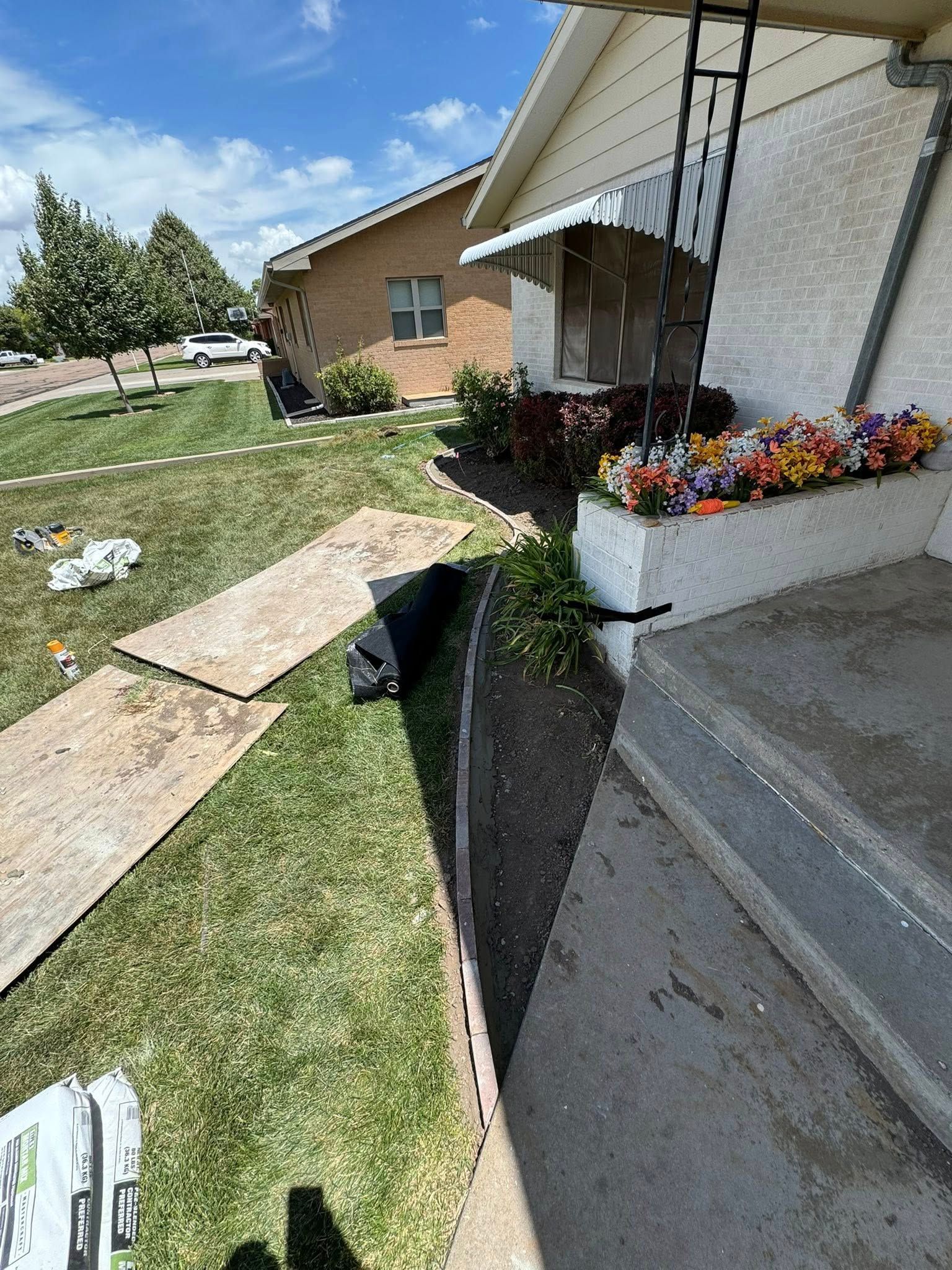 Damaged sidewalk beside a house with a deep trench, plywood boards, and flower planter near the porch
