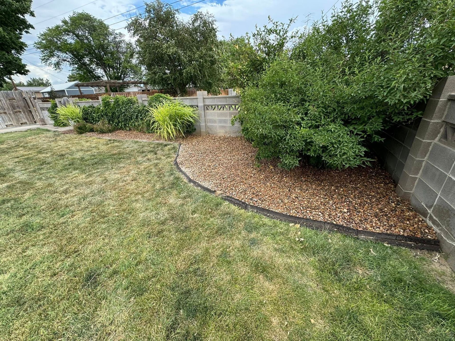 Backyard garden with grass, mulch bed, shrubs, and a stone wall under a cloudy sky