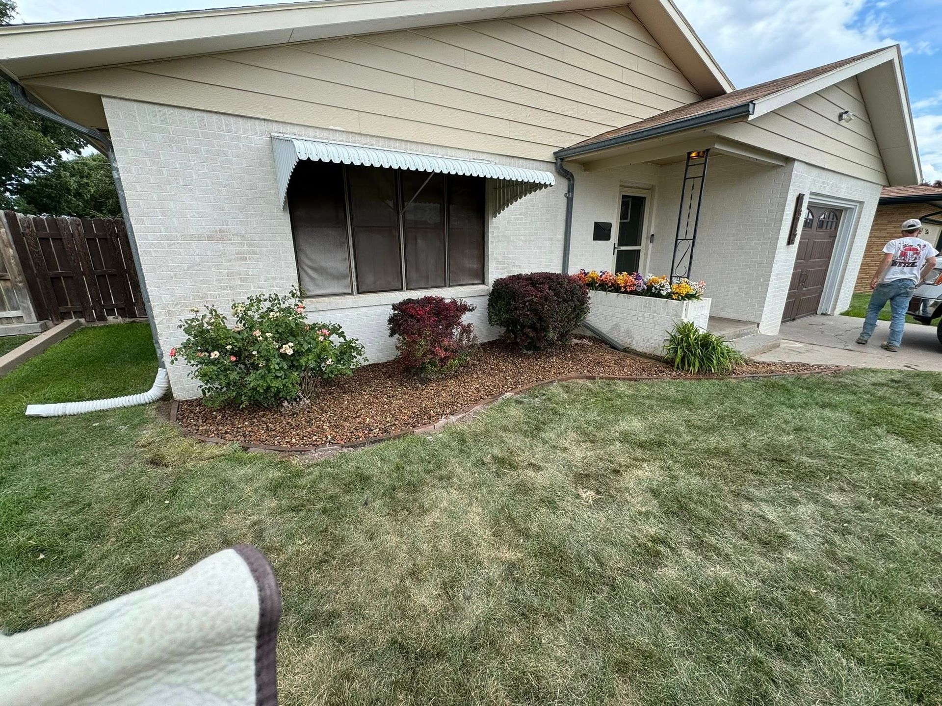 Suburban house exterior with white siding, front porch, shrubs, and a person watering plants on the right