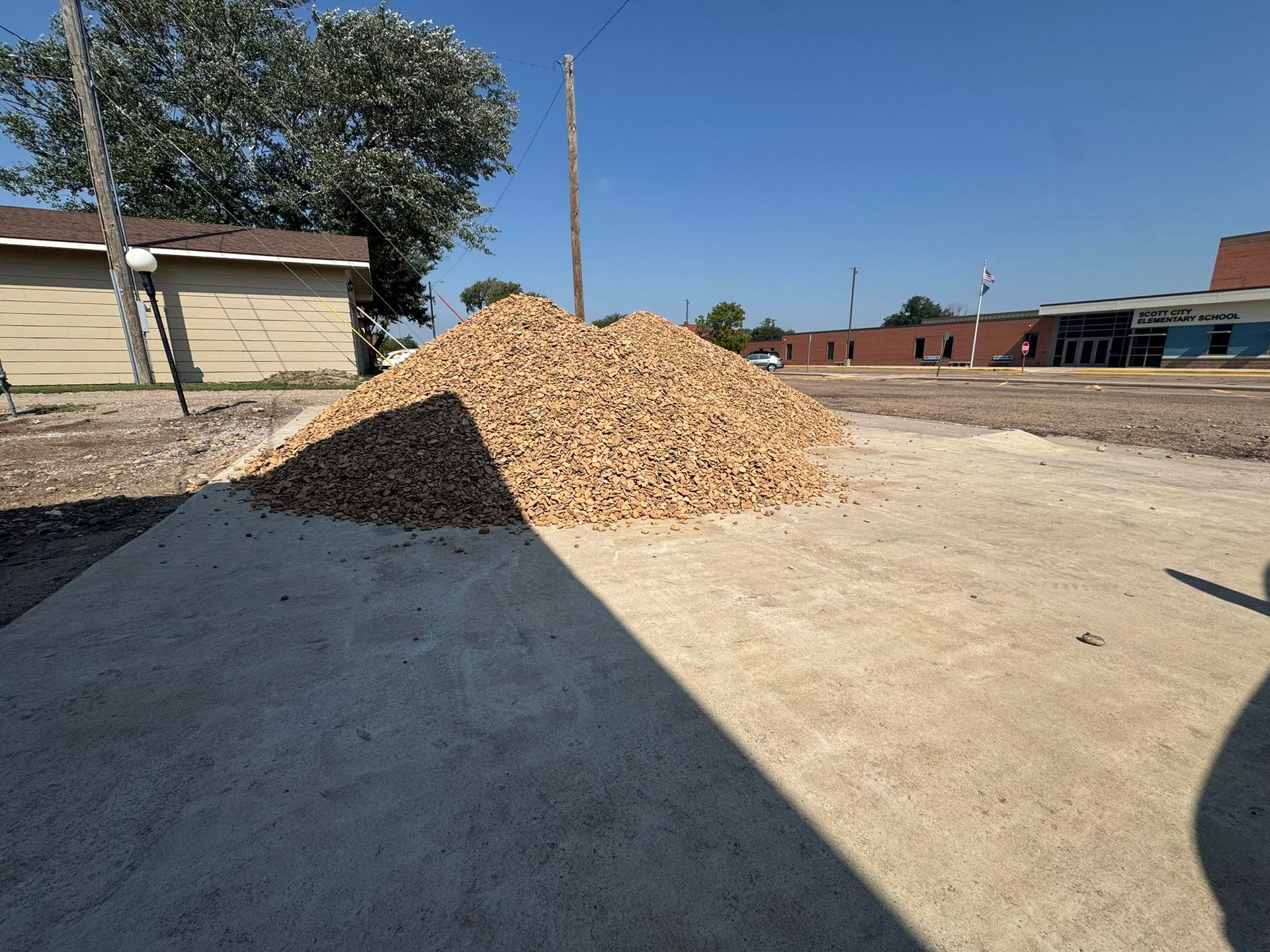 Large pile of wood chips on a sunny outdoor lot beside buildings and a road