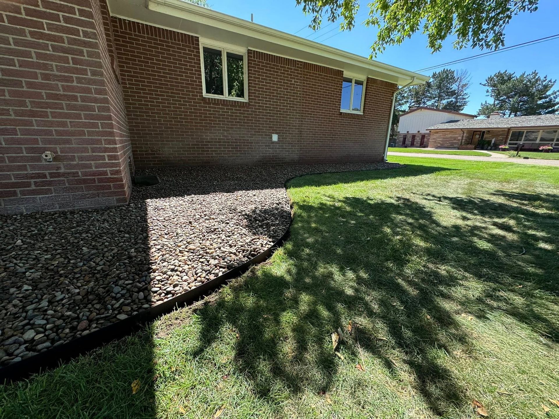 Brick house exterior with a window, mulch bed, and sunlit grass casting tree shadows