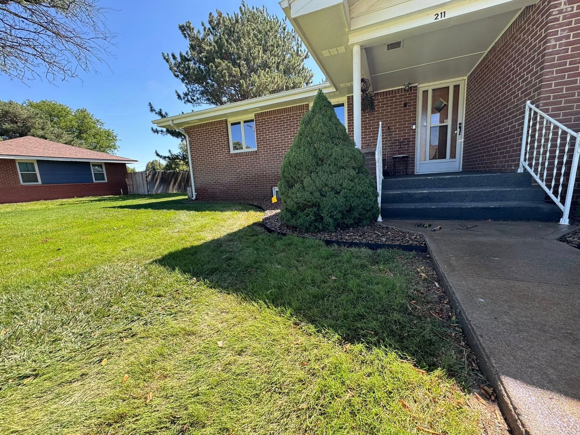Brick house front porch with concrete steps, white railing, and a green lawn in sunlight