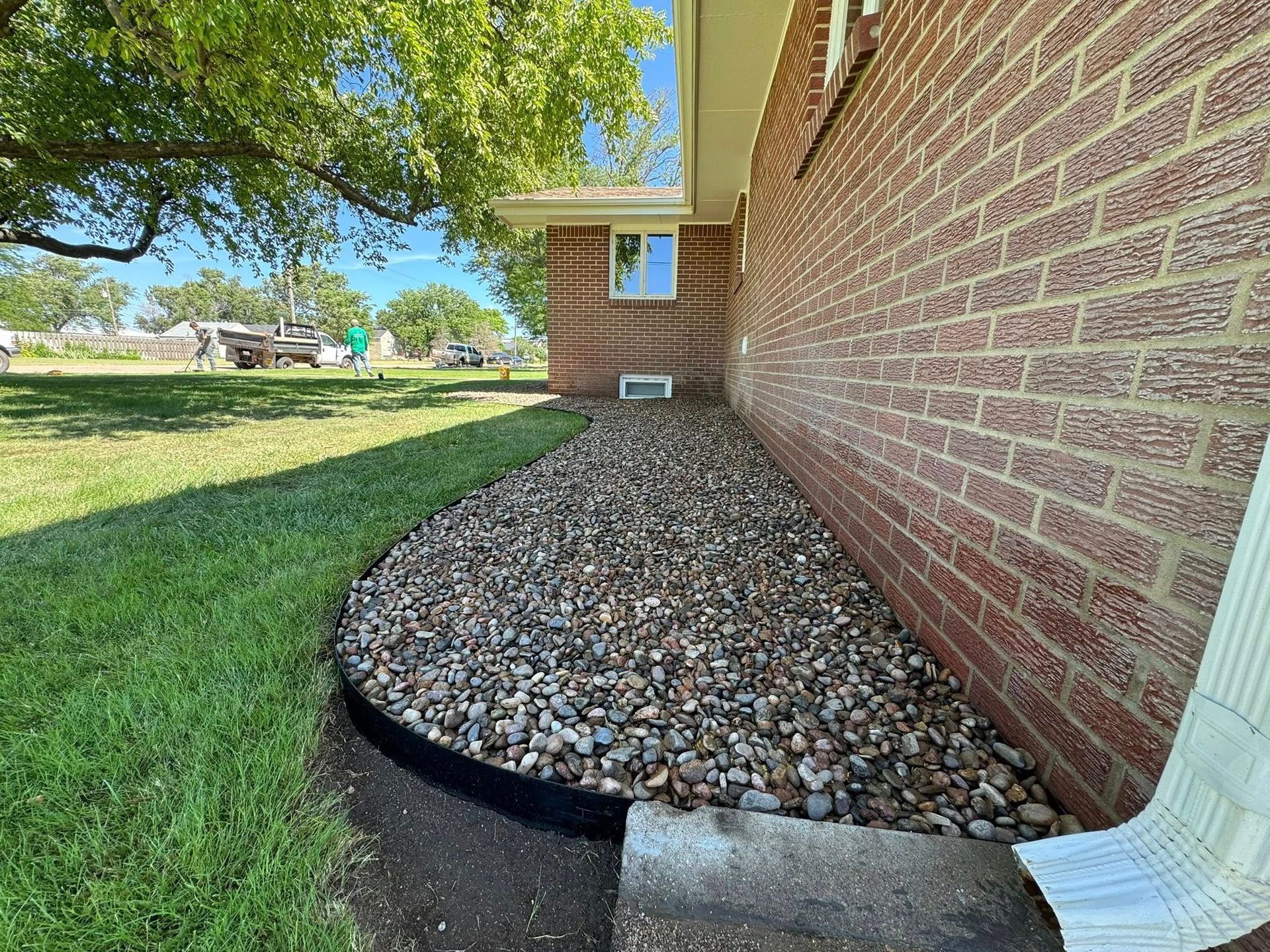Curved rock bed along a brick house wall beside a green lawn, with white downspout and window.