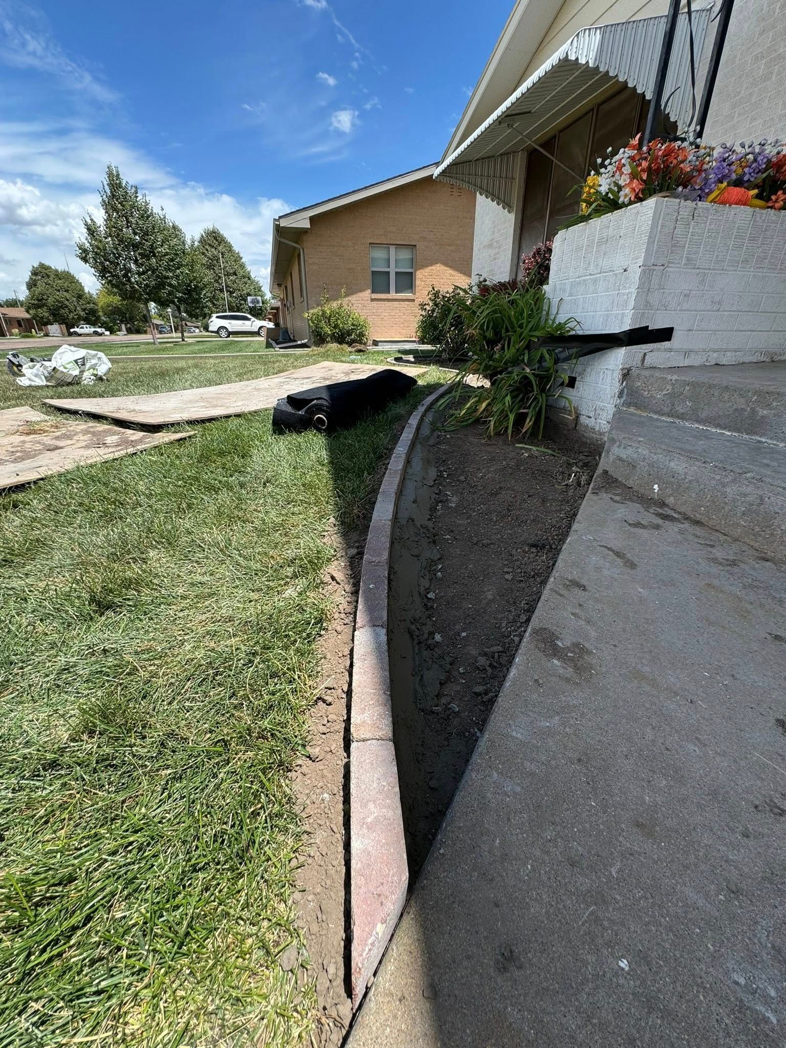 Residential sidewalk and driveway beside a brick house, with a small tree, lawn, and hanging flowers under a bright sky.