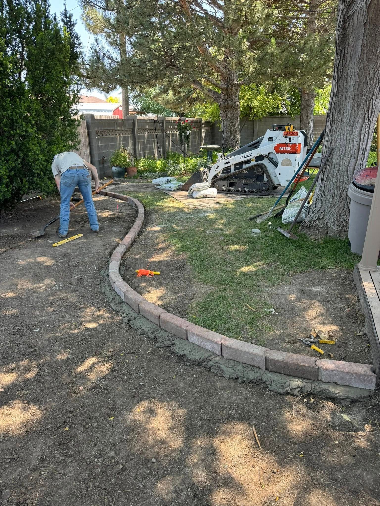 Worker trimming bushes along a curved garden border beside a white utility vehicle in a shady yard