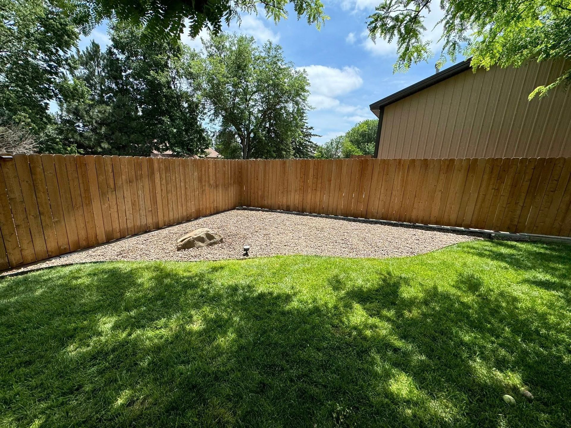 Backyard with wooden fence, gravel area, and green grass under a sunny sky