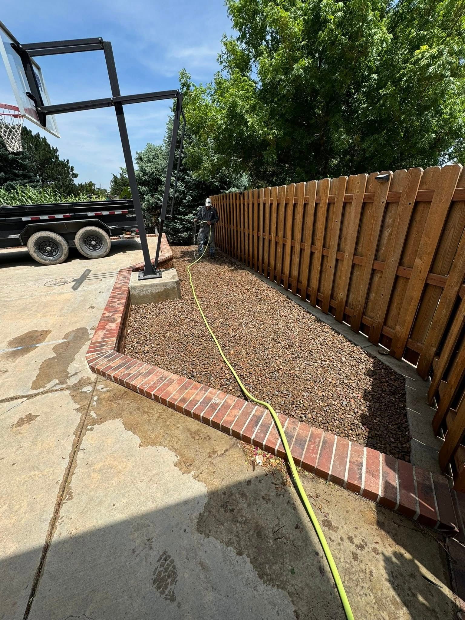Wooden fence beside a gravel yard with a brick border, measuring tape, and parked trailer on a sunny day