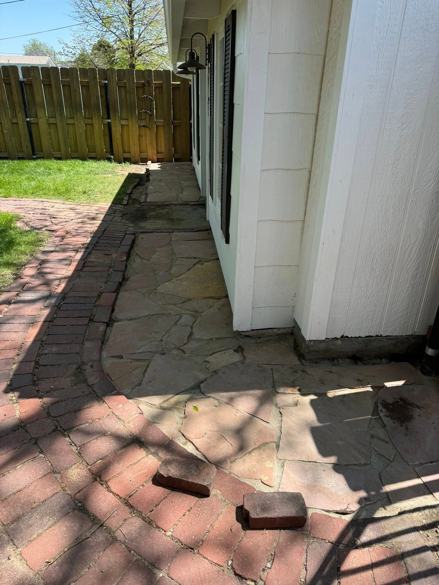 Narrow brick walkway beside a white house, with a wooden fence and lawn in sunlight.