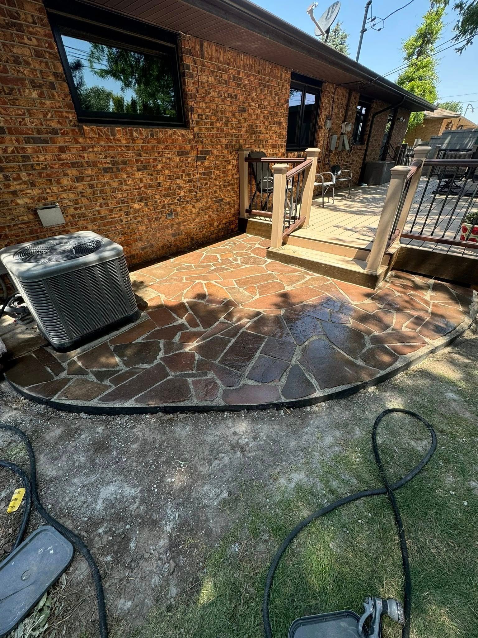 Brick patio beside house with wet stone surface, railing, outdoor unit, and garden hose in foreground