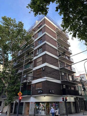 Esquina de la calle con un alto edificio de apartamentos de ladrillo y tiendas en la planta baja bajo un cielo azul.