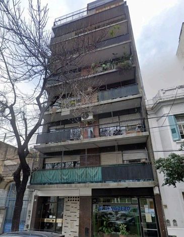Edificio de apartamentos alto y estrecho con balcones y plantas, visto desde la calle junto a árboles sin hojas.