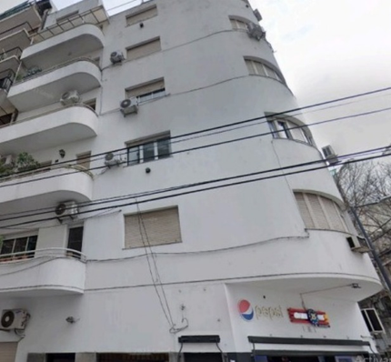 Edificio de apartamentos alto y blanco con balcones y letreros de tiendas a nivel de calle bajo un cielo nublado.
