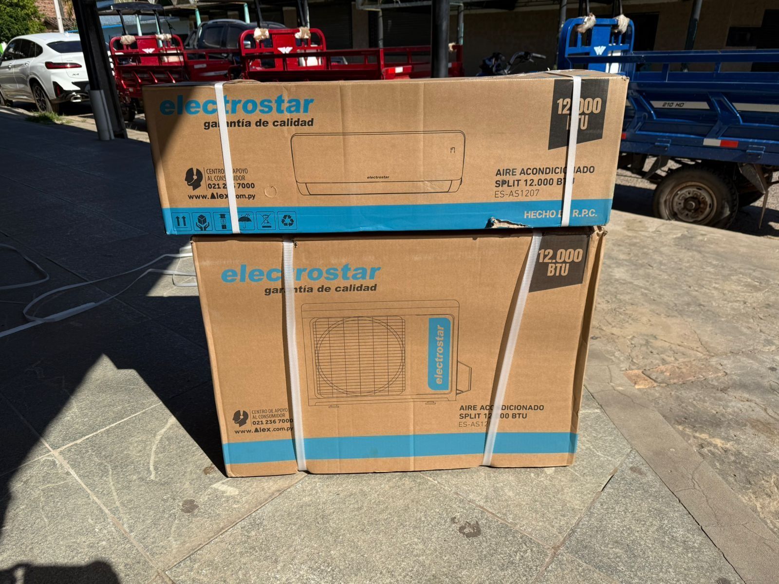 Dos cajas de cartón de Elecrostarr apiladas en un terreno de hormigón soleado cerca de camiones rojos y azules.
