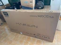 Una gran caja de cartón para televisor con la etiqueta “Mazon” y “Powered by webOS Hub” sobre un suelo de baldosas.