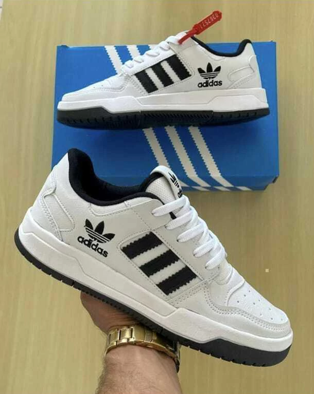 Zapatillas blancas estilo Adidas con rayas negras, una mostrada sobre una caja azul y la otra sostenida en la mano.