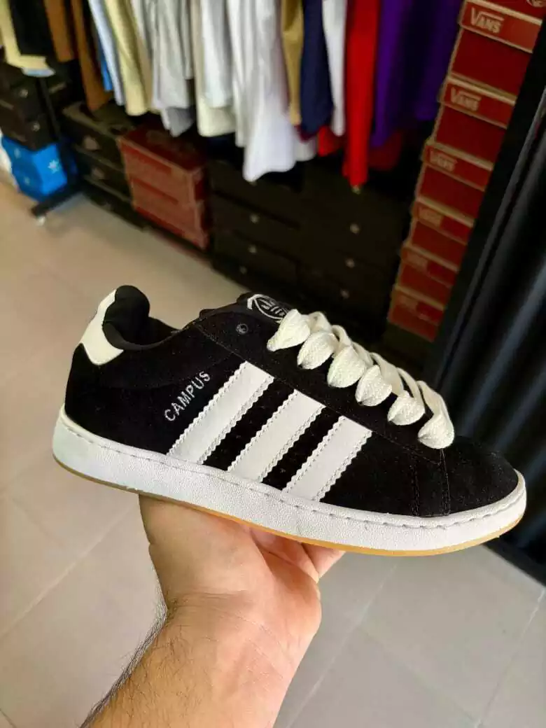 Una zapatilla Adidas negra con rayas blancas sostenida en una mano en el interior de una casa, con percheros de ropa al fondo.