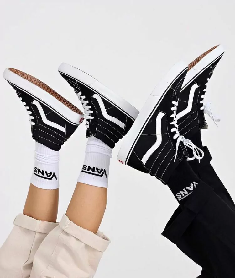Tres personas con las piernas levantadas, calzando zapatillas Vans blancas y negras y calcetines blancos sobre un fondo blanco.