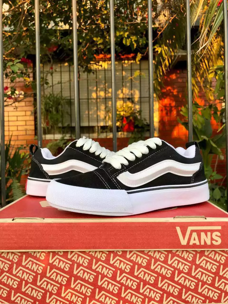 Zapatillas Vans blancas y negras sobre una caja roja de Vans al aire libre con plantas al fondo.