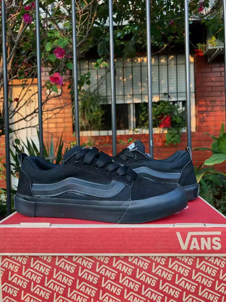 Zapatillas deportivas negras sobre una caja de zapatos Vans roja al aire libre, con una valla de jardín al fondo.