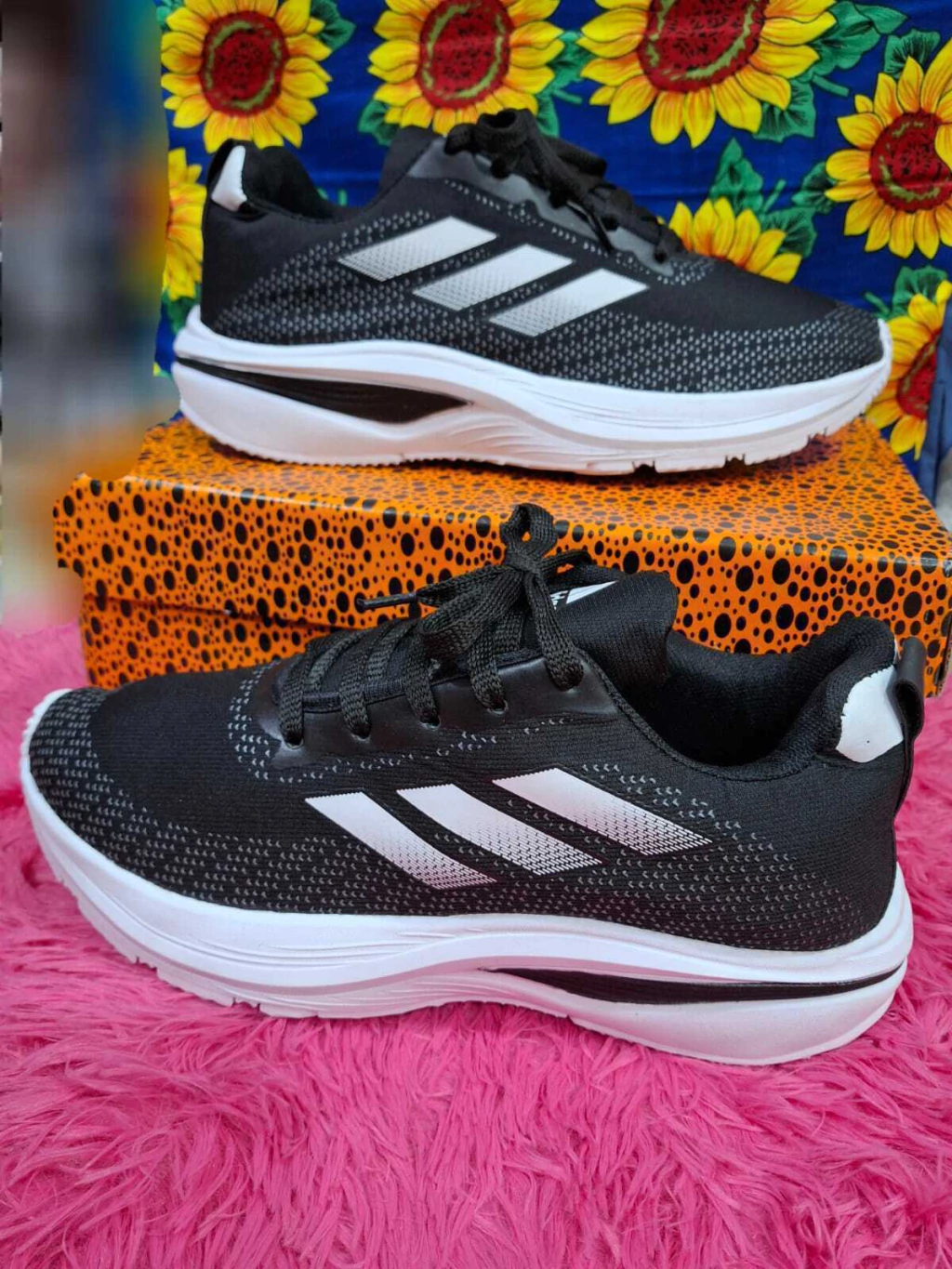Un par de zapatillas Adidas negras sobre pelo rosa, con una caja naranja con estampado de leopardo y flores de colores detrás.