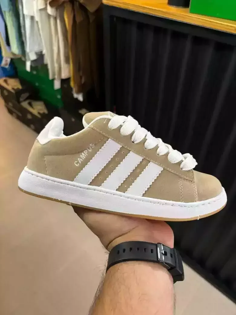 Una zapatilla Adidas color beige con rayas blancas, sostenida en el pasillo de una tienda.