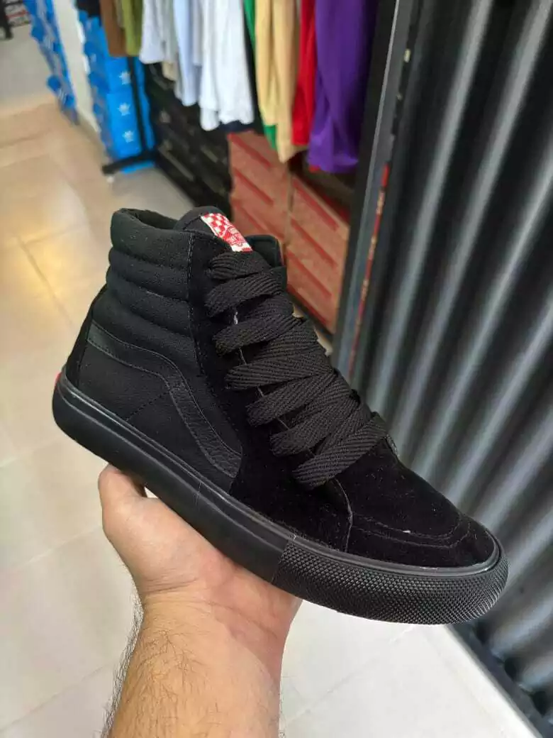 Una zapatilla deportiva negra de caña alta sostenida en la mano en el pasillo de una tienda, con filas de ropa al fondo.