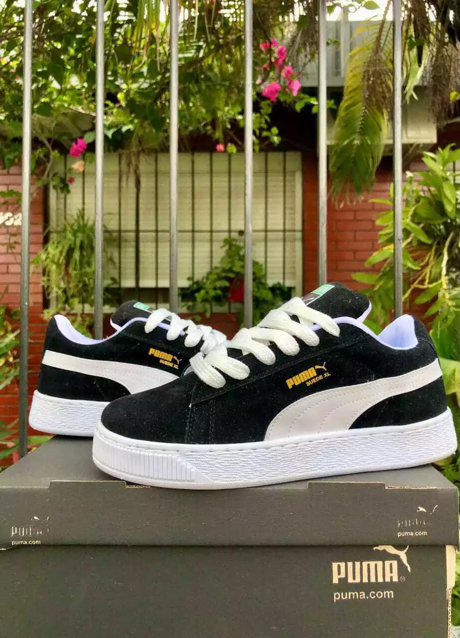 Zapatillas Puma negras con suela blanca sobre una caja al aire libre, con vegetación de fondo.