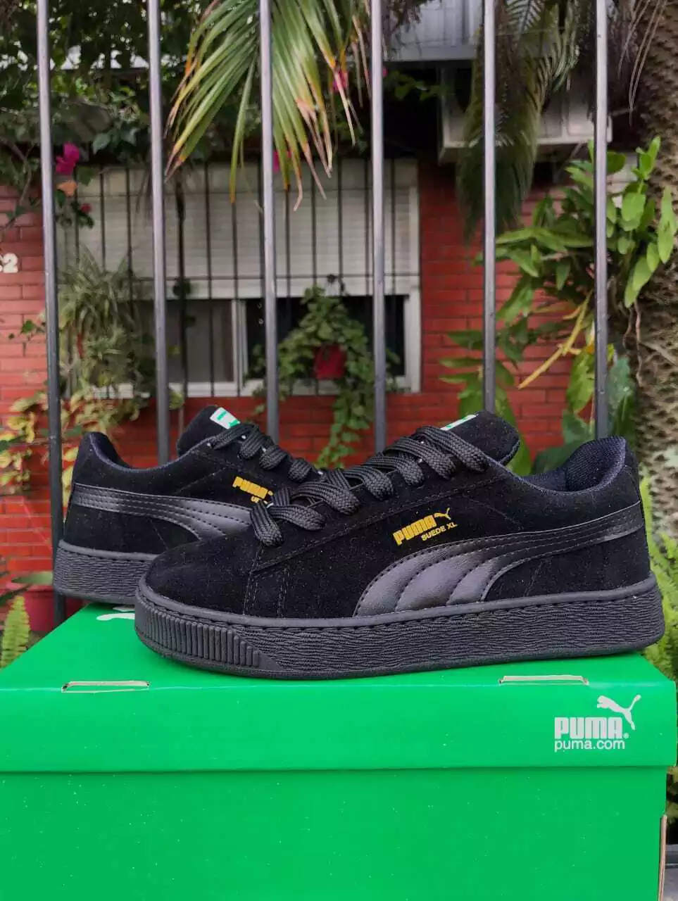 Zapatillas Puma negras sobre una caja verde al aire libre, con una pared de ladrillos y plantas al fondo.
