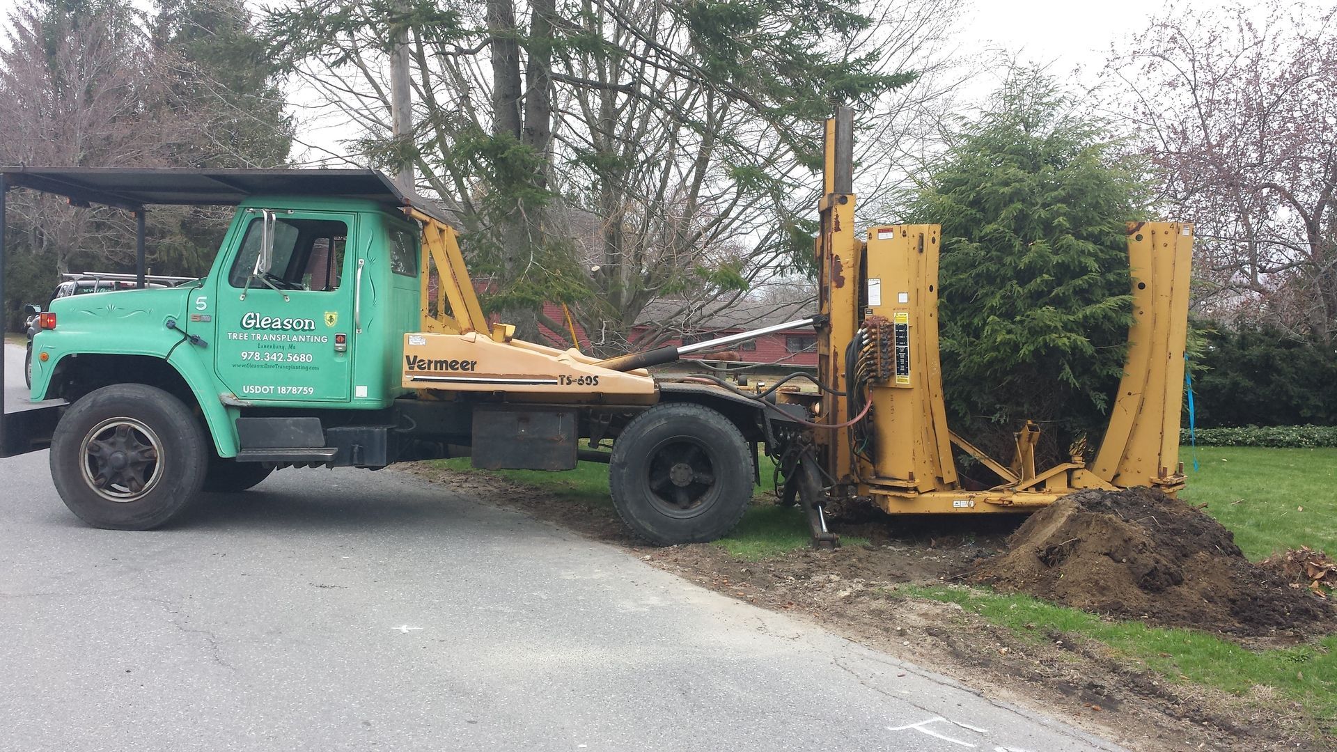 Green tow truck lifting a yellow hydraulic attachment on a roadside beside trees