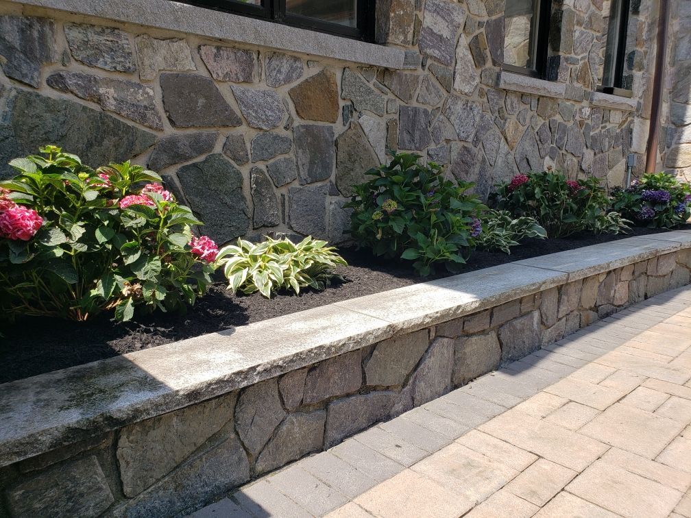 Stone planter bench along a sidewalk beside a stone wall, with pink and white flowers and green shrubs.