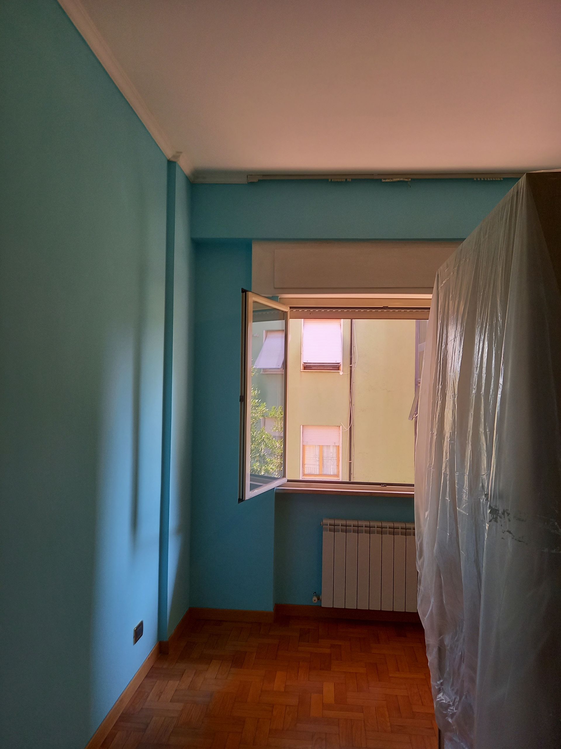 Stanza vuota con pareti blu, pavimento in legno e una finestra che si affaccia su un altro edificio.