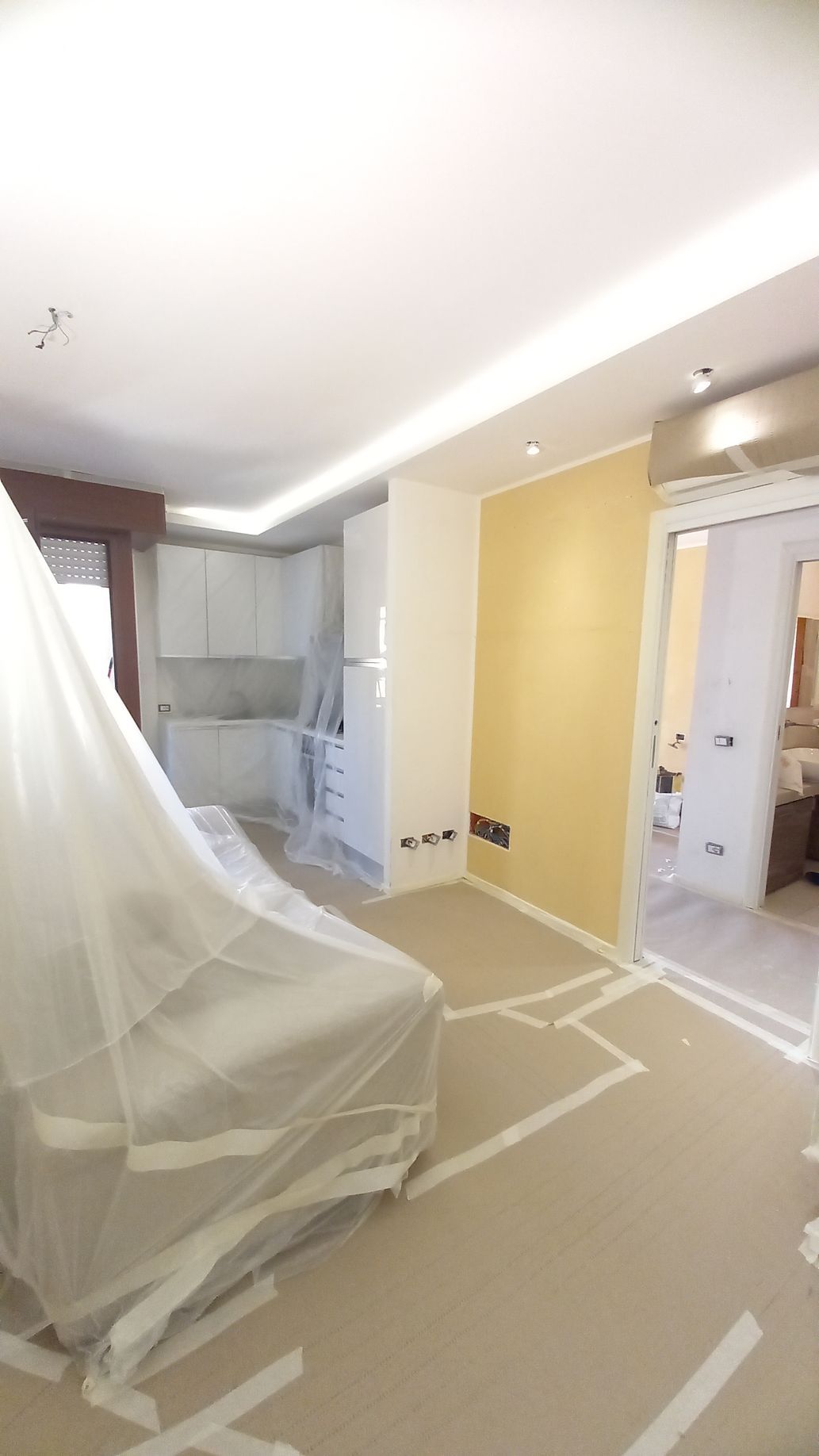 Stanza in ristrutturazione con mobili ricoperti di plastica, linee del pavimento tracciate con nastro adesivo e pareti gialle sotto luci intense del soffitto.