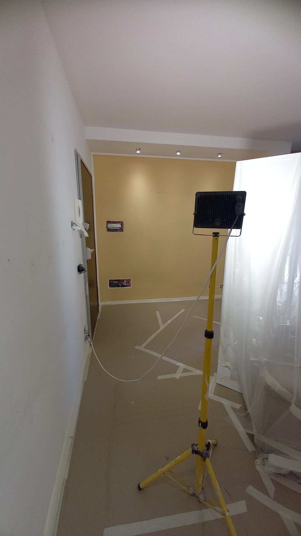Lampada da lavoro gialla su treppiede in un corridoio in fase di ristrutturazione, con teli di plastica e rivestimenti del pavimento fissati con nastro adesivo.