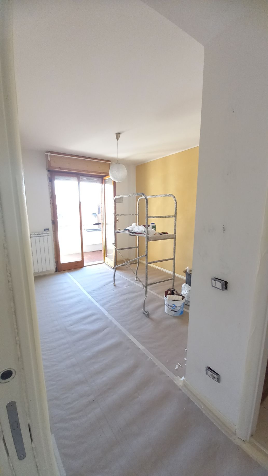 Stanza parzialmente ristrutturata con pareti bianche, pavimento grezzo e una piccola impalcatura vicino a una finestra luminosa.