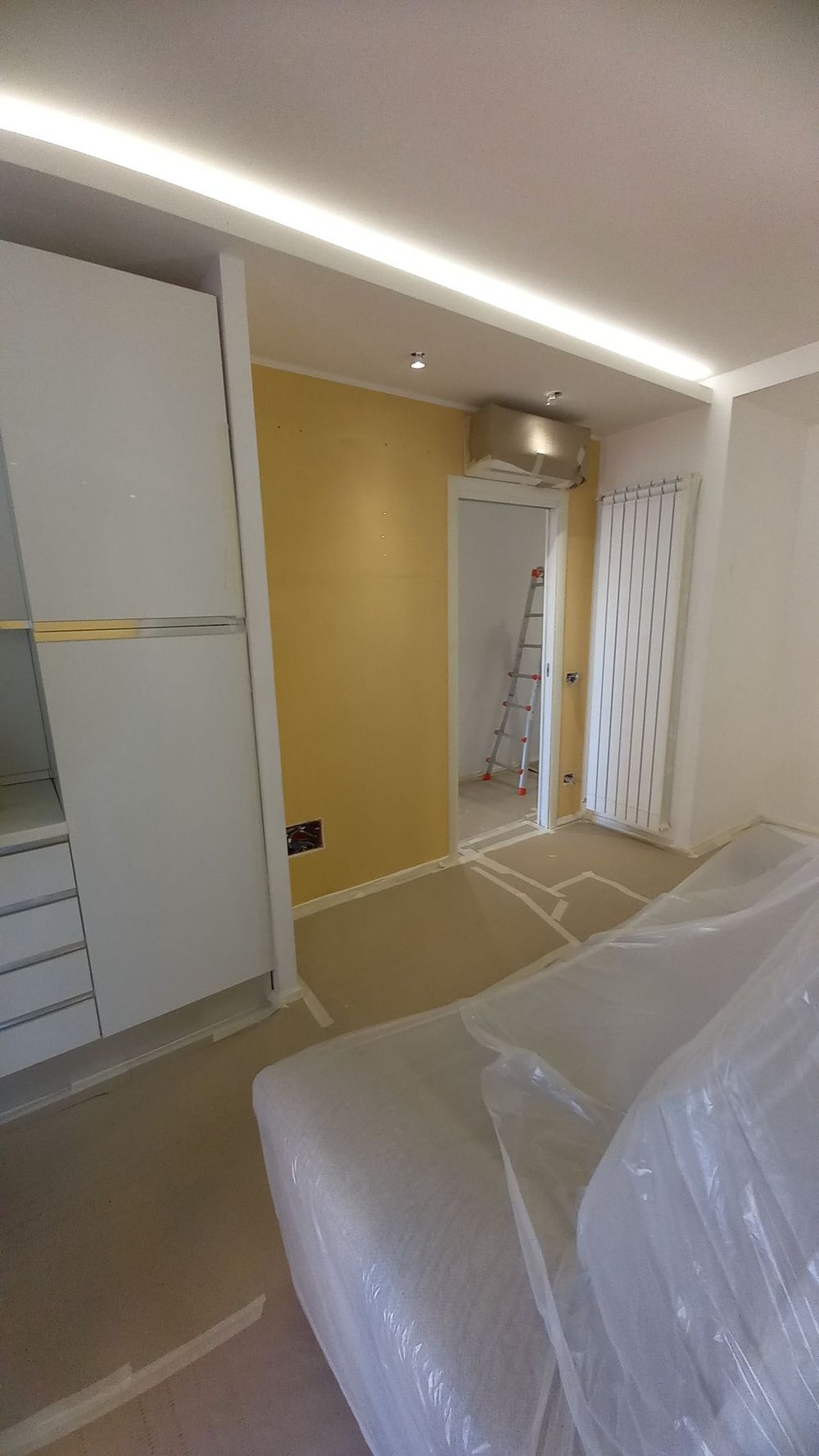 Camera da letto parzialmente arredata con armadio, parete decorativa gialla e mobili rivestiti in plastica.