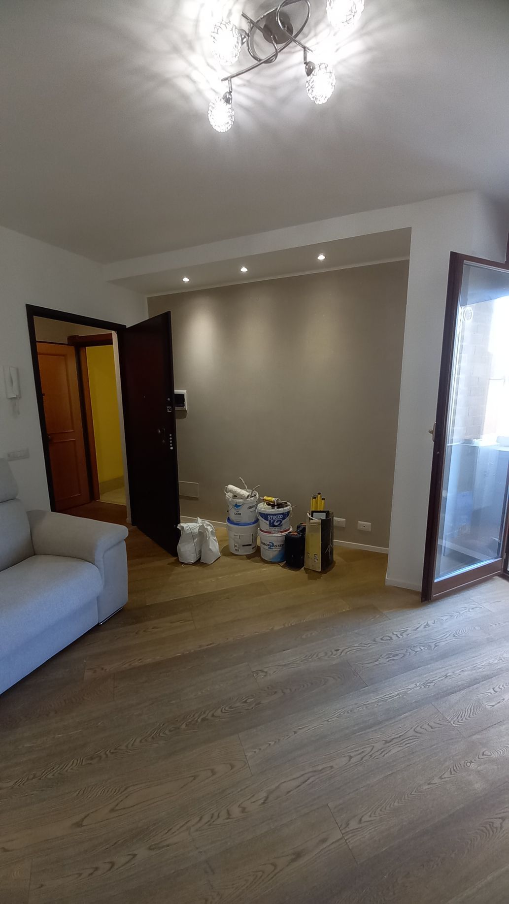 Soggiorno moderno con pavimento in legno, divano grigio, faretti a incasso e scatole vicino a una porta aperta.