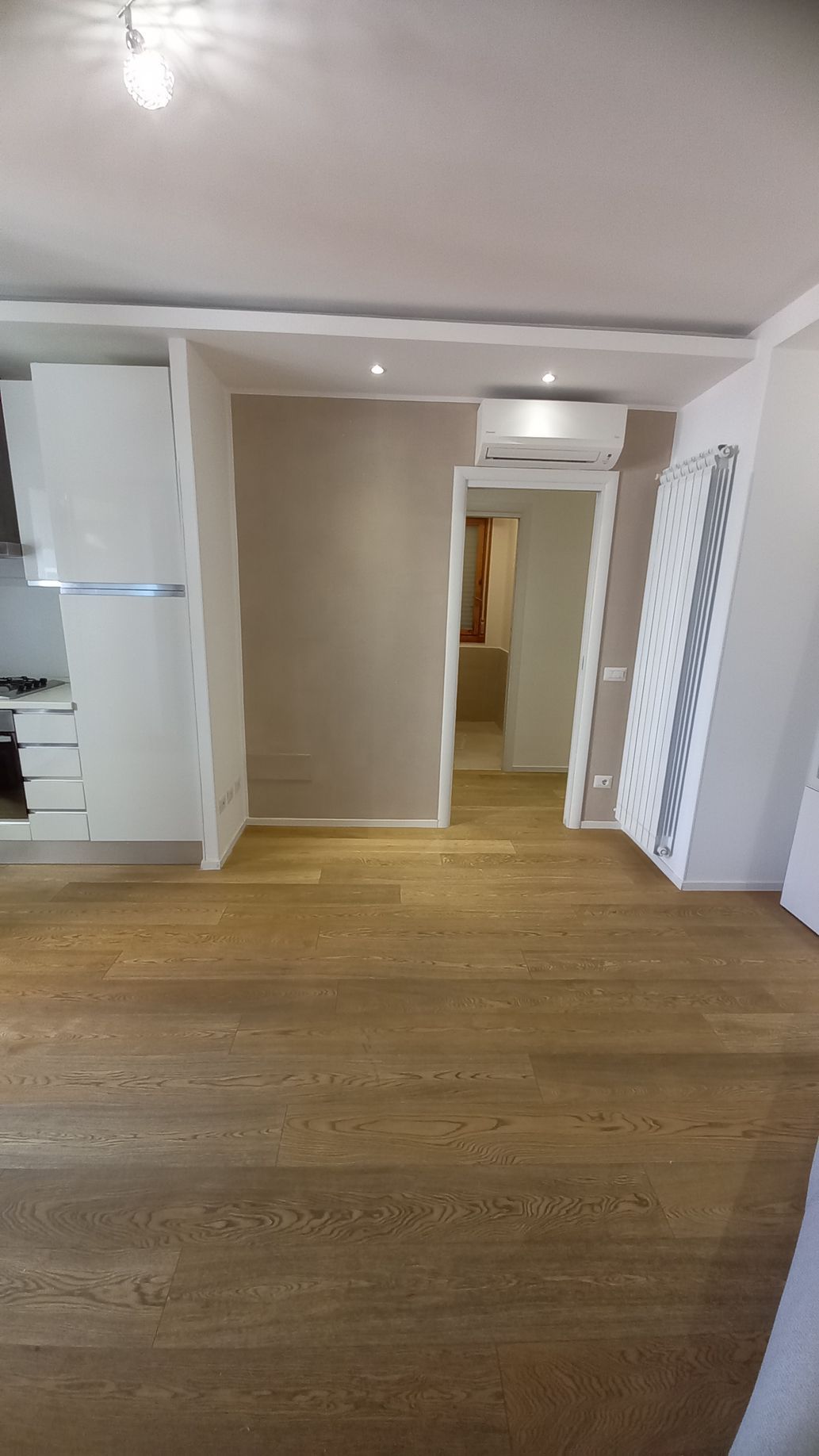 Soggiorno vuoto con pavimento in legno, pareti beige e porta che dà su un corridoio.