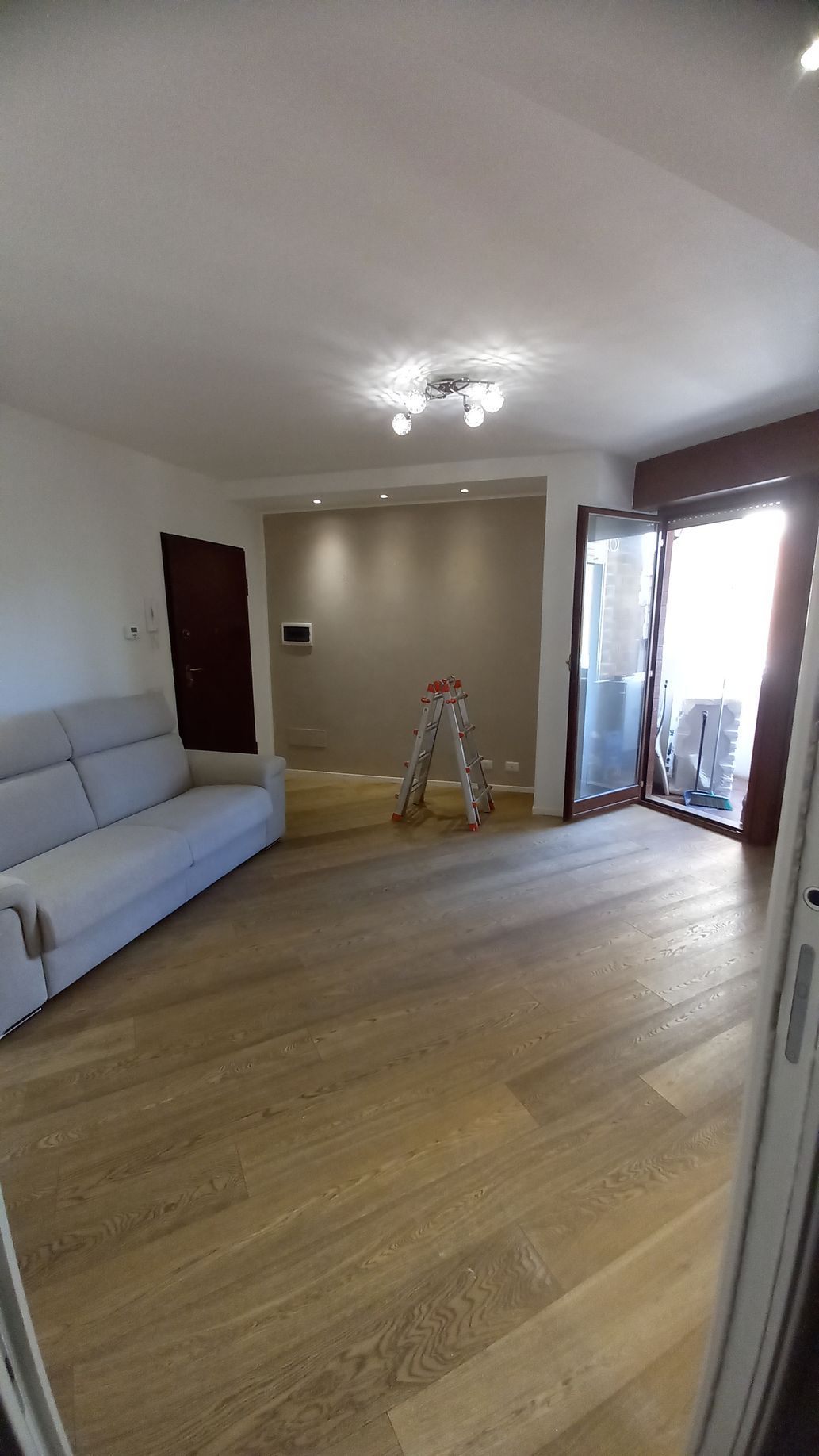 Luminoso soggiorno con divano grigio, pavimento in legno, porta finestra che si apre sul balcone e una scaletta a pioli vicino alla parete.