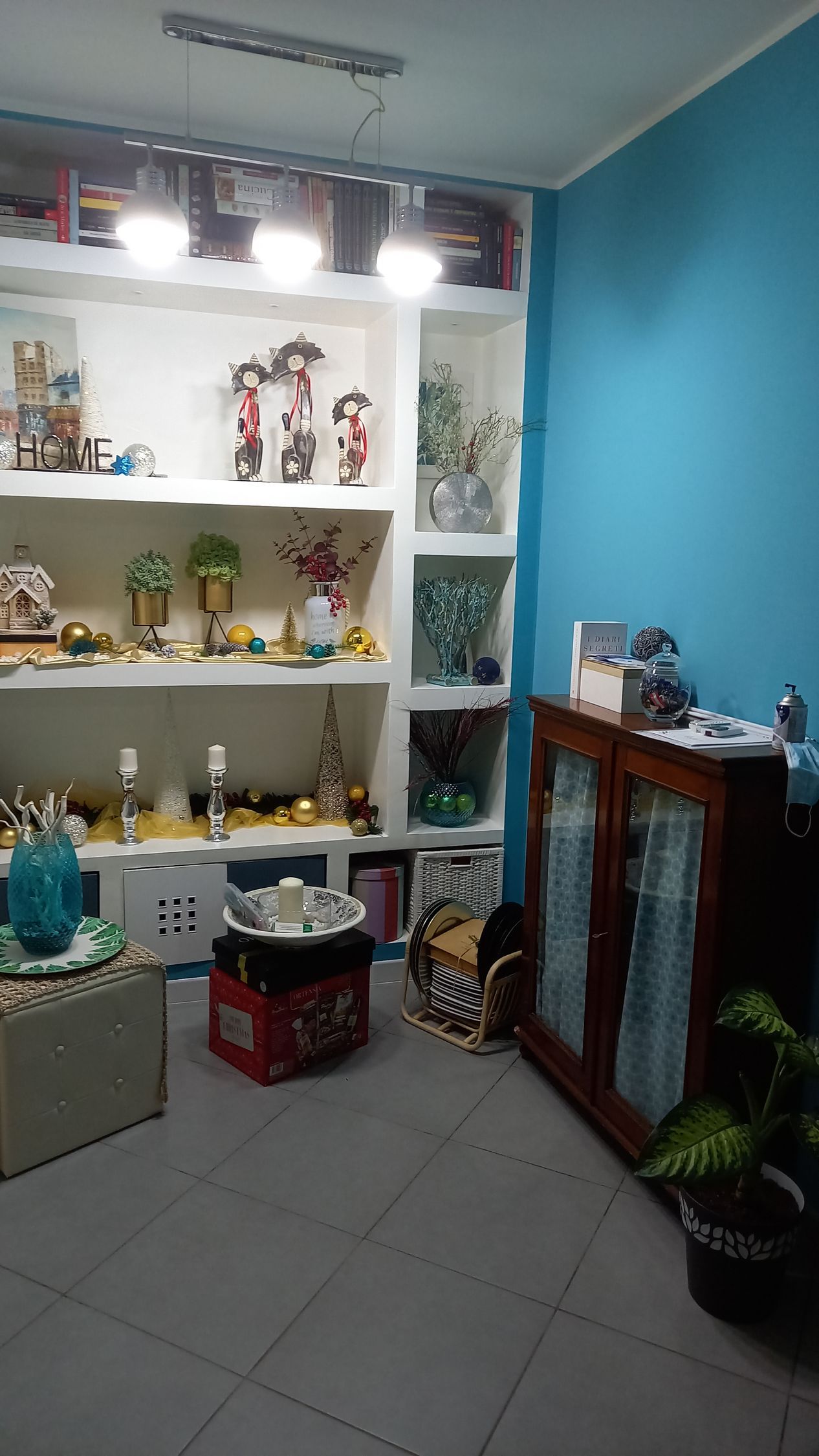Piccola vetrina con scaffali pieni di statuette, una parete blu e oggetti decorativi sui tavoli.