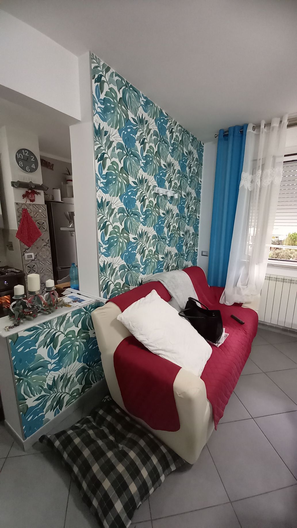 Luminoso soggiorno con divano rosso e bianco, parete decorata con motivi floreali, tende blu e pavimento piastrellato.