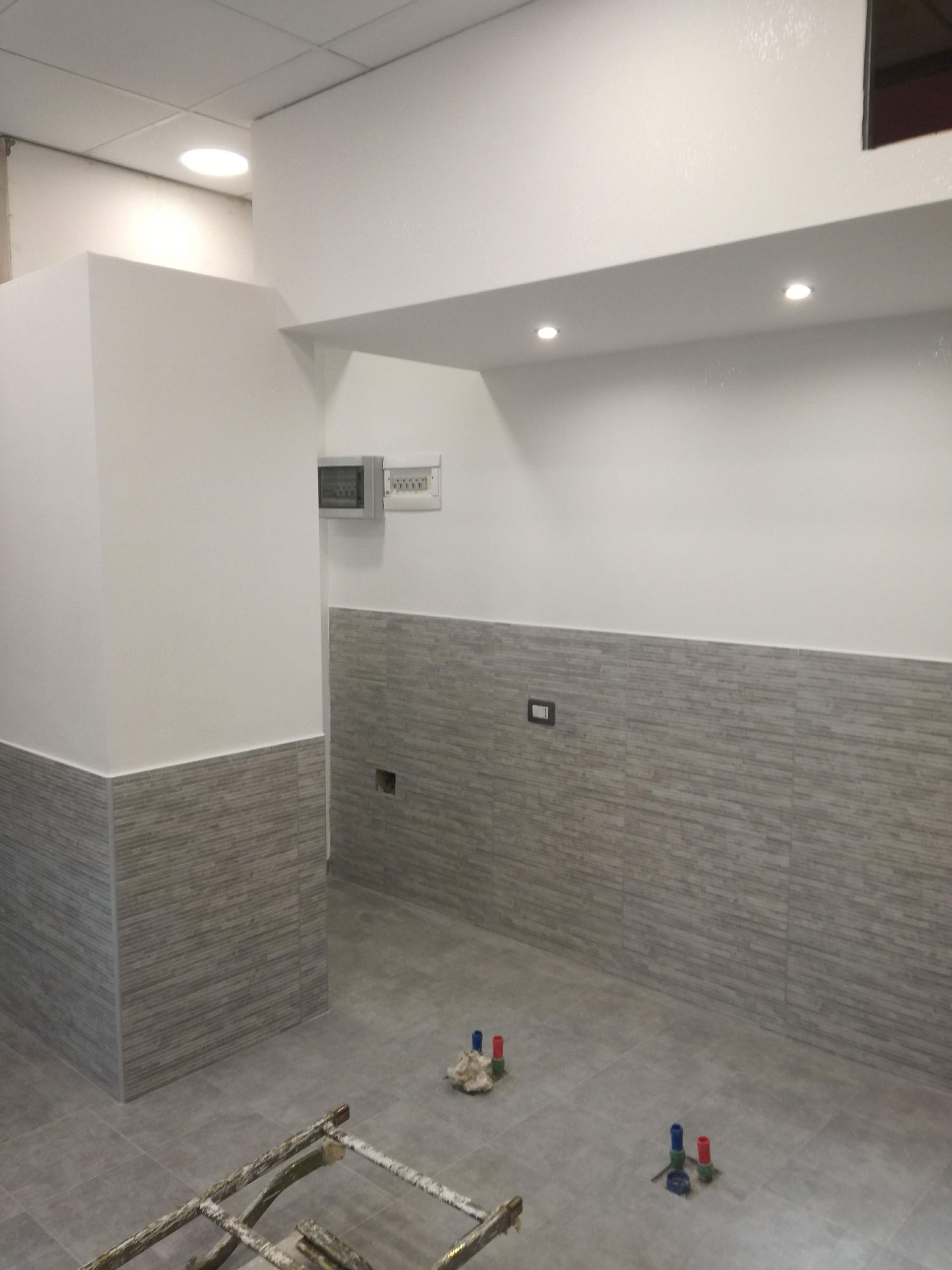 Stanza vuota piastrellata in fase di ristrutturazione con piastrelle grigie alle pareti, faretti a soffitto e rubinetteria sul pavimento.