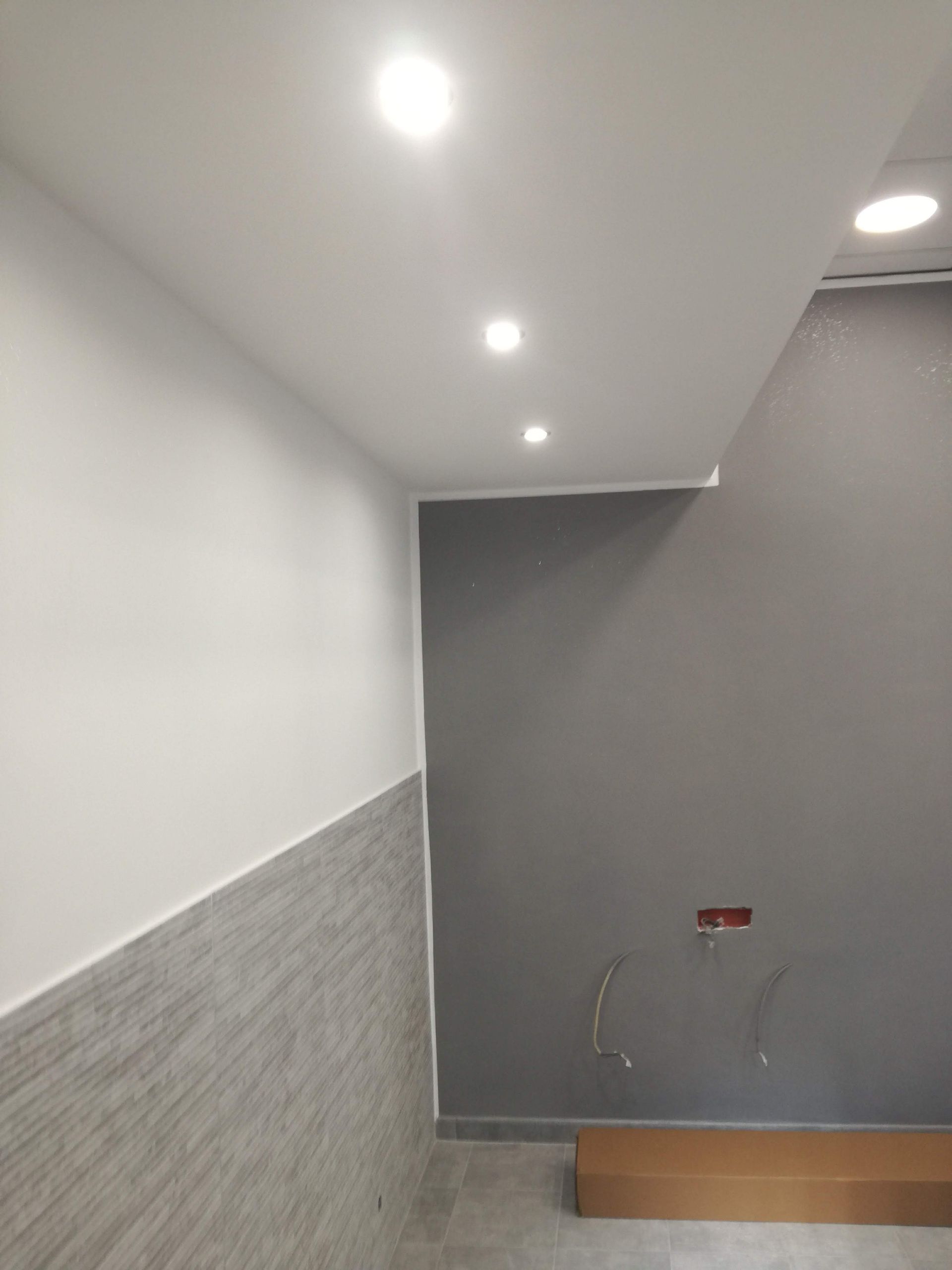 Stanza vuota con lampade a soffitto bianche, pareti grigie e una piccola zona di servizio non rifinita.