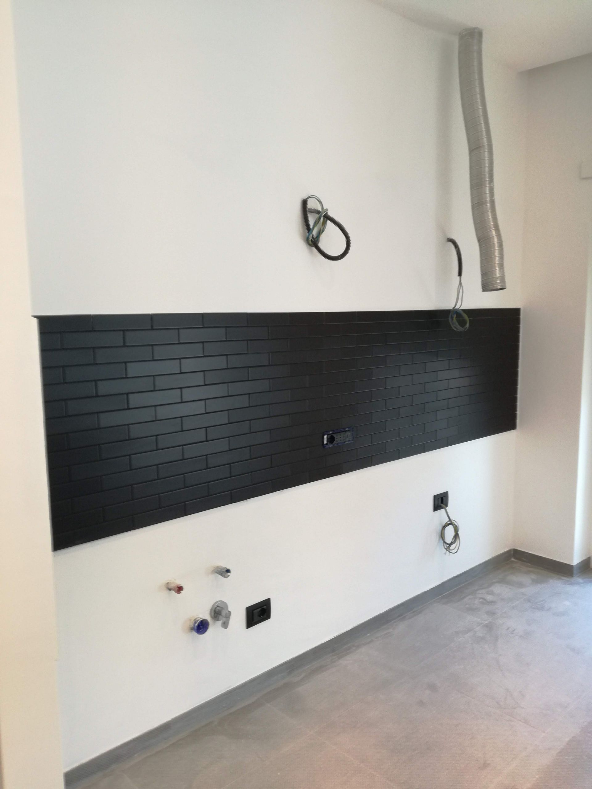 Parete della cucina non rifinita con rivestimento in piastrelle nere, prese elettriche a vista e pavimento grigio.